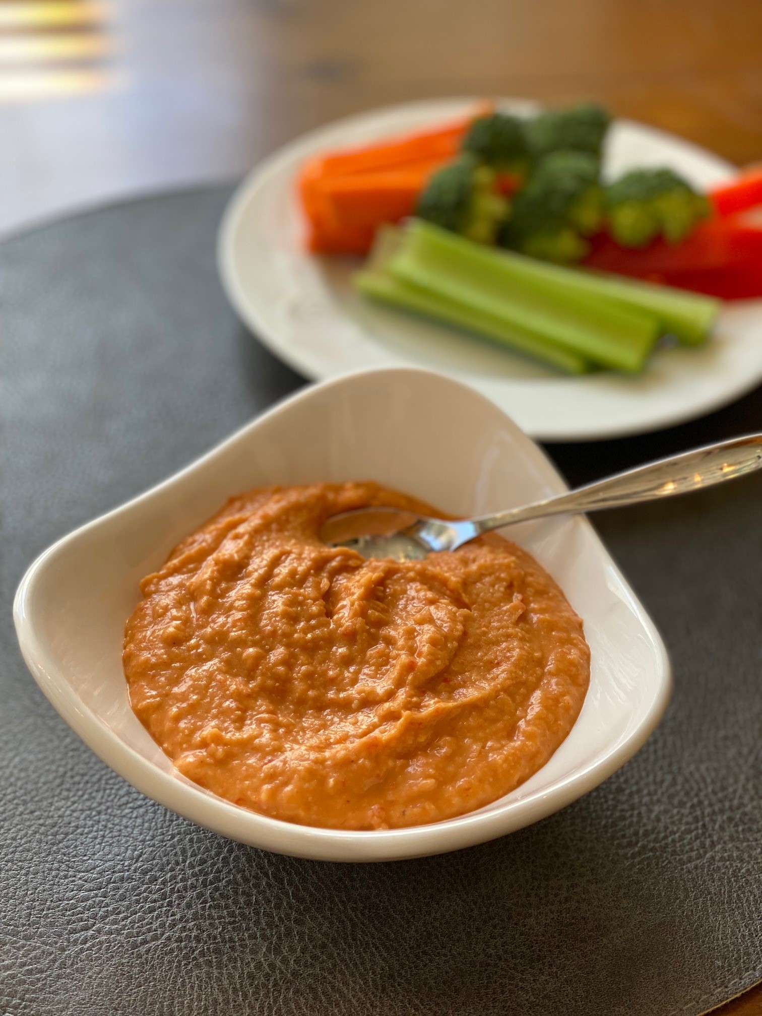 a bowl of red pepper dip in front of a plate of raw vegetables