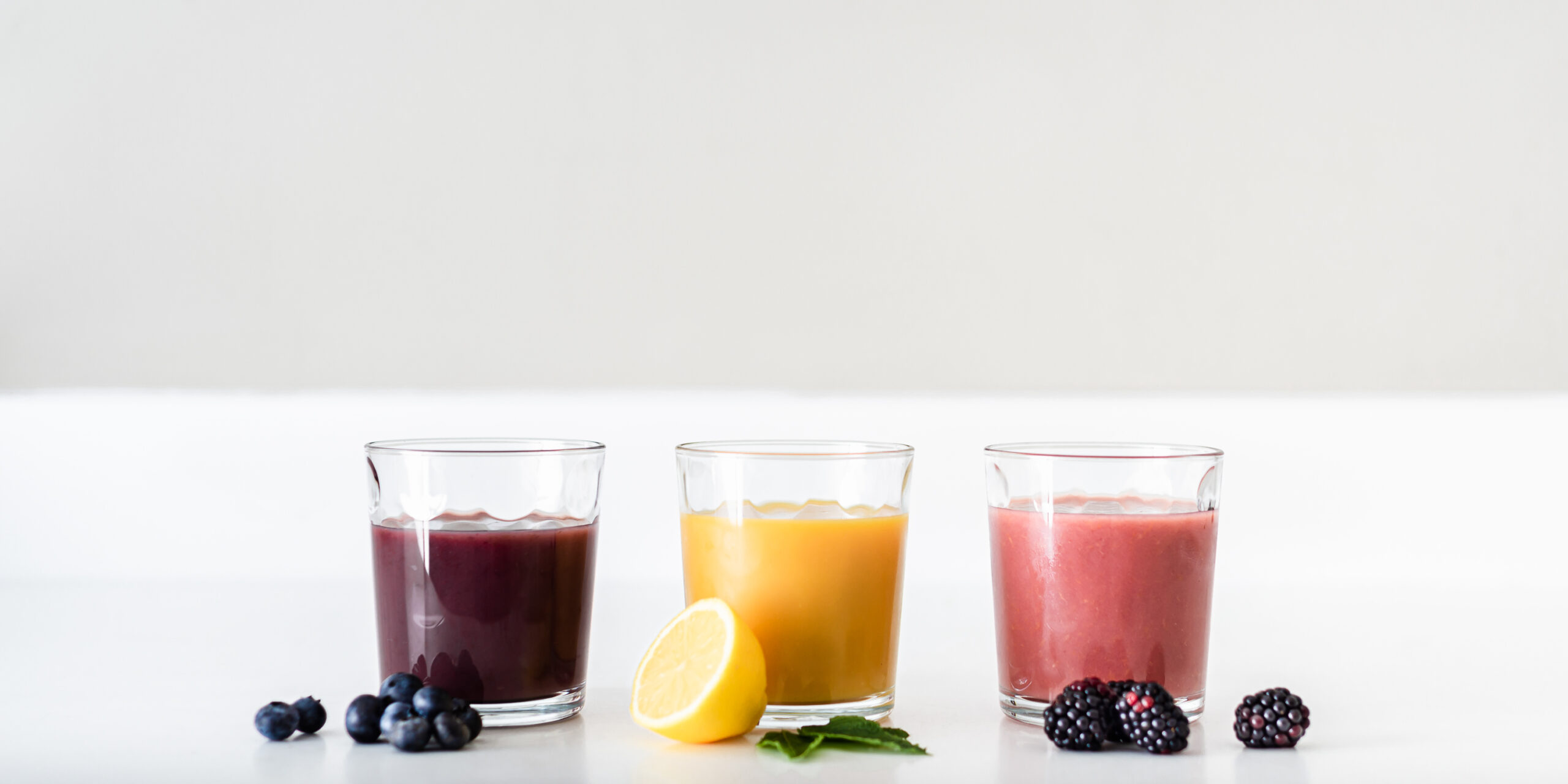 A row of three juice glasses with three different colored juices and some fruit., representing probiotics and prebiotics.