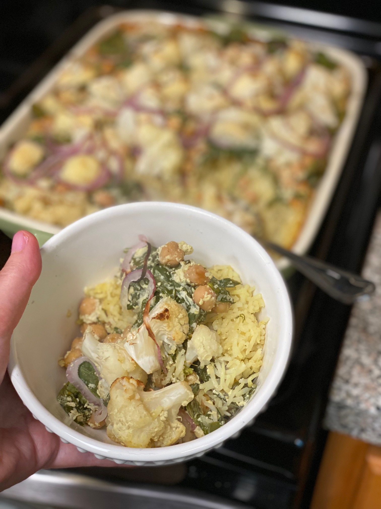 A person holding a bowl of a cauliflower and rice dish in front of the casserole.
