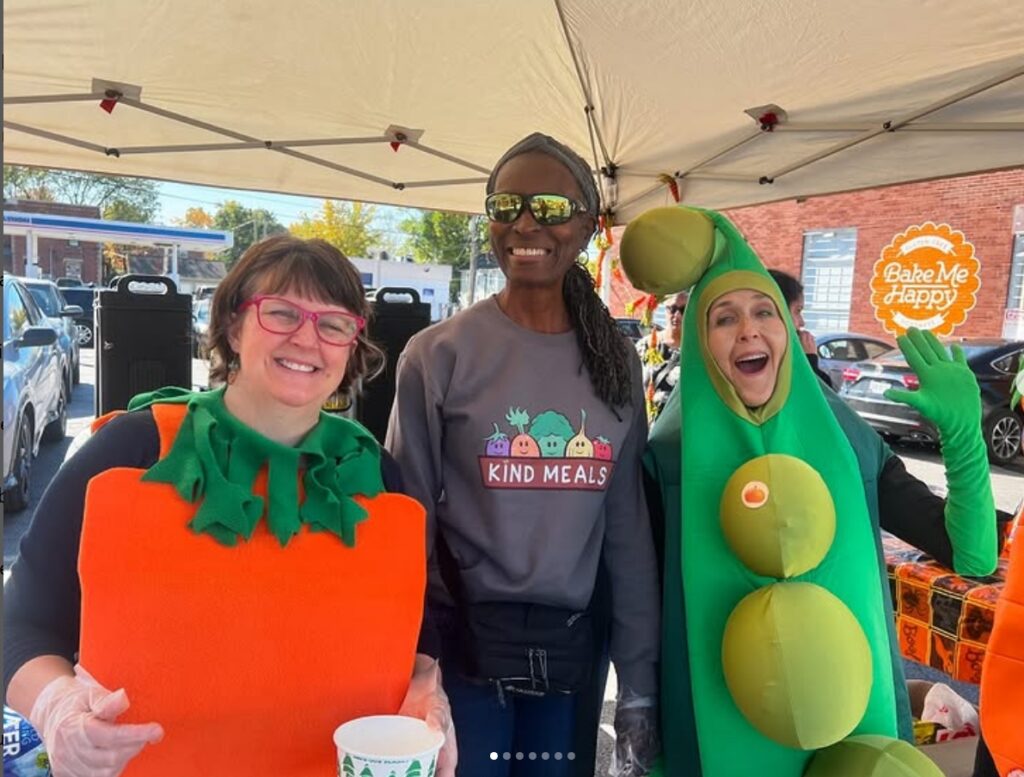 Three smiling people - one is dressed as a carrot and one is dressed in a peapod costume, the third is wearing a sweatshirt with the Kind Meals logo.